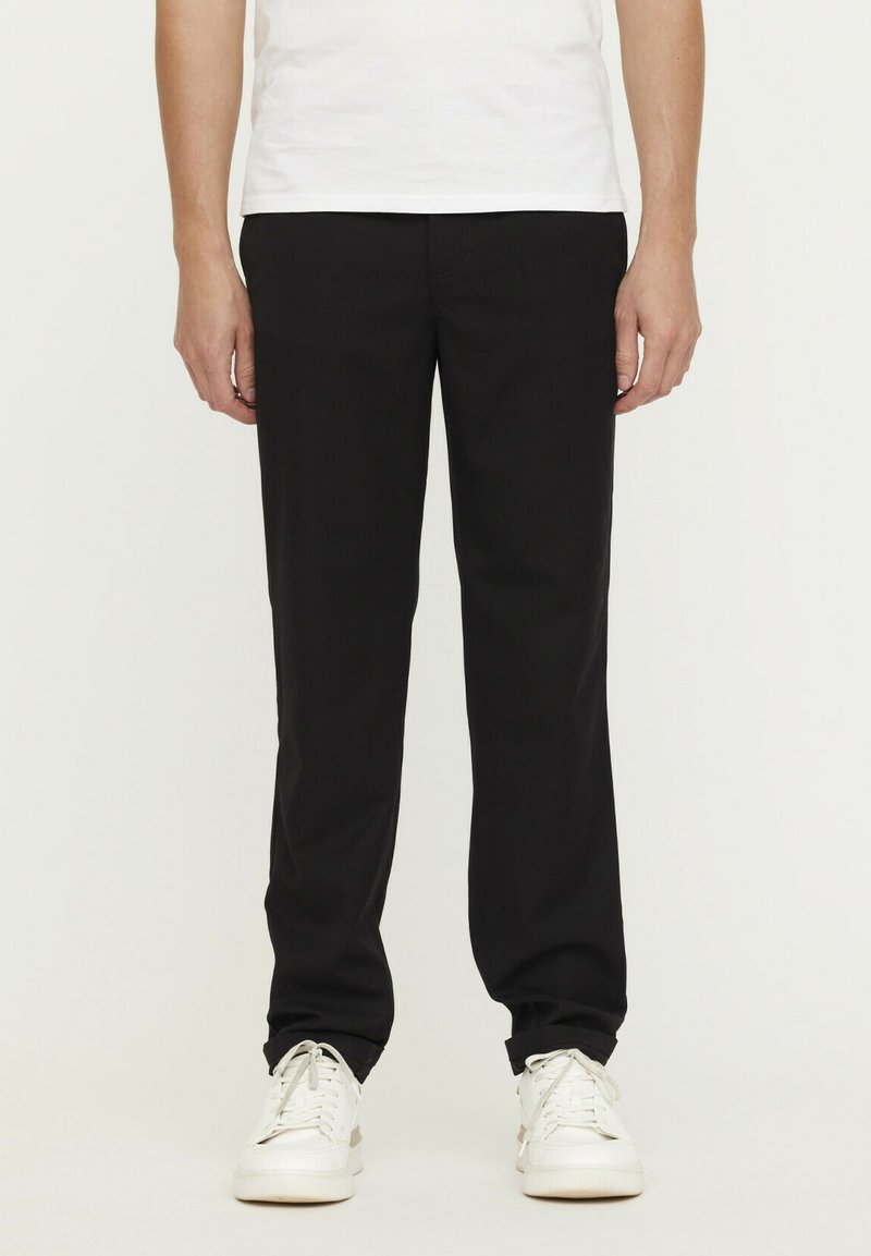 Pantalon noir en tissu tissé, avec une coupe droite, des poches avant et une texture lisse, associé à des baskets blanches.