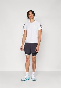 Camiseta blanca con logo y franjas negras de Adidas en las mangas, combinada con pantalones cortos negros y pantalones cortos negros debajo, zapatillas blancas con detalles en azul.