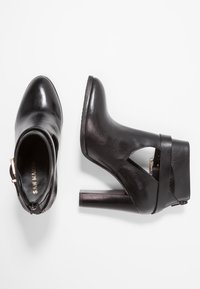 Musta nahkasaapas nilkkapituudella, teräväkärkinen, leikattu sivuilta, kultaiset yksityiskohdat ja tukeva korko. Sileä pinta ja yksinkertainen muotoilu.