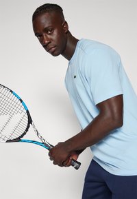 T-shirt azzurro chiaro a maniche corte con scollo rotondo, con un logo verde. Il modello tiene una racchetta da tennis nera e blu.
