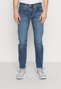 Jeans de mezclilla azul con un ajuste entallado, con una cintura de altura media, cinco bolsillos y costuras de contraste. Combinados con zapatillas grises.