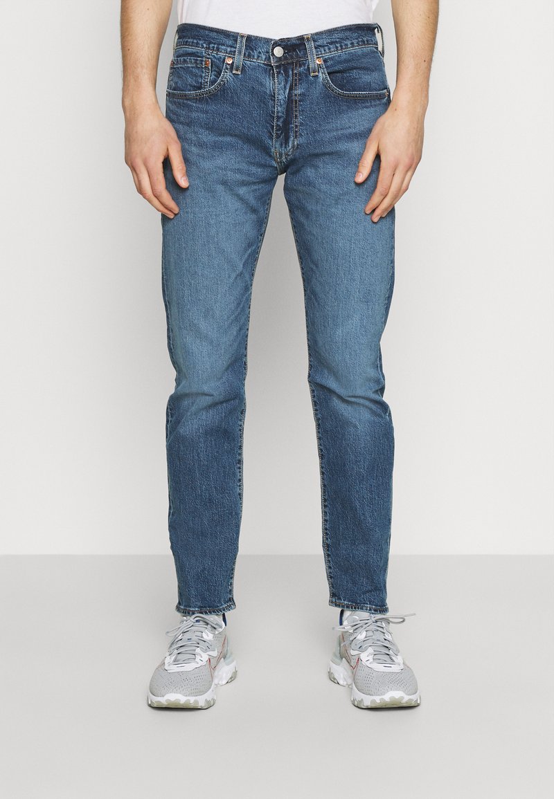 Jeans de mezclilla azul con un ajuste entallado, con una cintura de altura media, cinco bolsillos y costuras de contraste. Combinados con zapatillas grises.