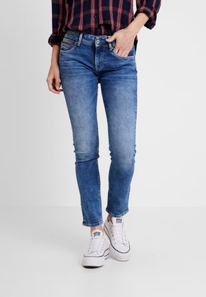 Person, der schmal geschnittene blaue Jeans, ein rot-navy karierte Hemd und weiße flache Sneaker trägt, steht mit einer Hand in der Tasche.