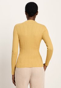 Person mit dunkel geflochtenem Haar im Dutt, der einen engen, gerippten senffarbenen Pullover und helle beige Hosen trägt, steht mit den Händen in den Taschen, Rückansicht.