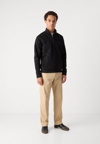 Pullover in fleece nero con collo a zip a quarti, caratterizzato da un design delle spalle cucito, abbinato a pantaloni beige e scarpe nere.