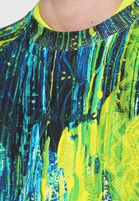 Rövid ujjú ing élénk kék-zöld absztrakt mintával, sárga díszítéssel, bordázott gallérral és hímzett logóval.