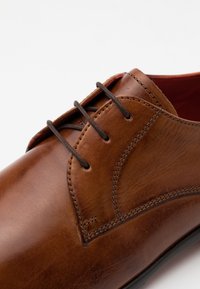 Zapato de vestir de cuero marrón con un acabado suave, que presenta una punta redondeada, cuatro cordones marrones y detalles de costura visibles a lo largo del costado.