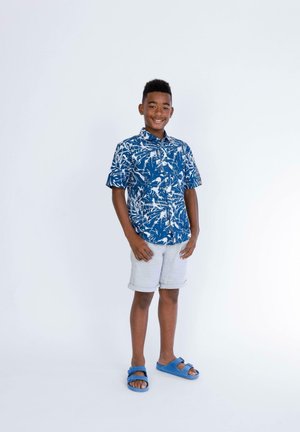 Chemise bleue à manches courtes avec un motif floral blanc, shorts gris clair et sandales bleues. Posture debout sur un fond uni.