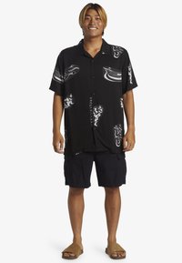 Camisa negra de manga corta con patrones gráficos en blanco, combinada con pantalones cortos cargo oscuros y sandalias de tiras beige. Ajuste casual y diseño relajado.