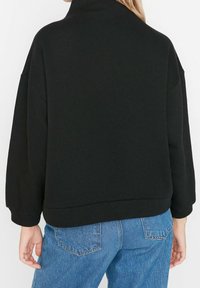 Sudadera negra con cuello alto, hombros caídos y puños de canalé. Combinada con jeans de tiro alto de color azul claro, mostrando un ajuste relajado.