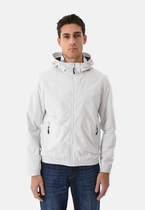 Uomo che indossa una giacca con cappuccio grigio chiaro con cerniere e tasche con cerniera, in piedi davanti a uno sfondo bianco uniforme.