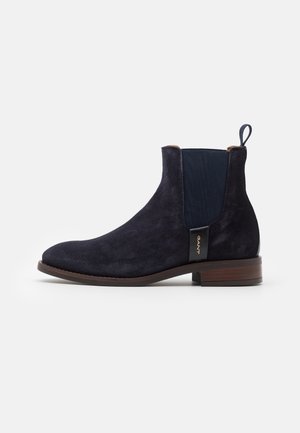 Bottine Chelsea en daim bleu marine avec panneau latéral élastique, languette de tirage, petit talon marron et étiquette en cuir brandée sur le côté.