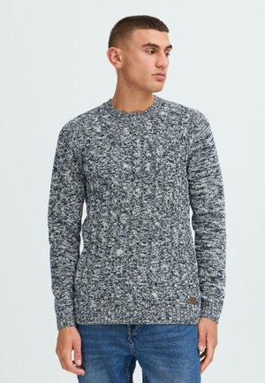 Knit sweater with a textured pattern in navy and white, crew neck, and long sleeves. Features a small logo patch on the lower side.