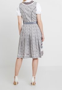 Kobieta stoi, ubrana w sukienkę w kwiatowy wzór sięgającą do kolan, z szarym pasem i puffowanymi białymi krótkimi rękawami, w połączeniu z białymi szpilkami.