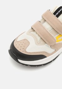 Adidași bej, albaștri și negri cu două benzi Velcro bej și un accent galben pe lateral, prezentați din unghiul din fața dreaptă.