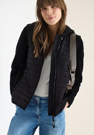 Jeune femme aux cheveux bruns portant un gilet matelassé noir sur des manches noires, une chemise claire, un jean bleu, et portant une sangle de sac à dos beige.
