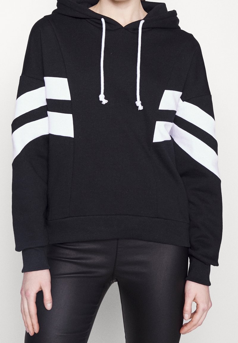 Personne portant un sweat à capuche noir avec des rayures diagonales blanches sur les manches et un pantalon en cuir noir, les mains détendues le long du corps.
