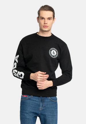 Junger Mann mit nach hinten gegelten Haaren, der einen schwarzen Pullover mit weißem Schriftzug am Ärmel und Logo auf der Brust trägt, kombiniert mit blauen Jeans.