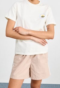 Persona che indossa una t-shirt bianca Adidas con logo e pantaloni corti beige chiaro, braccia incrociate, tatuaggio di un orso visibile sul braccio sinistro.
