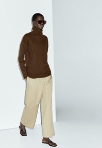 Brown turtleneck sweater, beige wide-leg trousers, and dark sunglasses. Smooth textures with a relaxed fit and minimalistic design.