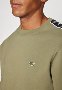 Sudadera verde oliva con cuello redondo, que presenta un pequeño logotipo de cocodrilo bordado y detalles en blanco y negro en los hombros.