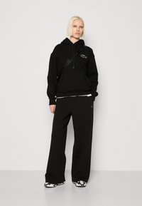 Felpa nera in cotone con tasca frontale, cappuccio con cordino e logo verde. Abbinata a pantaloni larghi neri e sneakers con motivo.