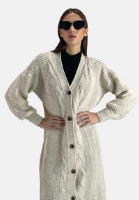 Cardigan lungo in maglia a trecce crema con un design testurizzato, grandi bottoni e maniche oversize; indossato sopra un top nero.