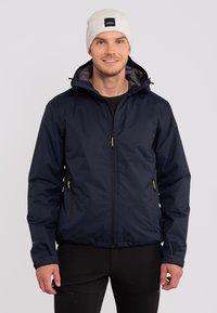 Marineblauwe jas met een ritssluiting aan de voorkant, twee zijzakken en een capuchon. Heeft gele accenten en een gladde, lichte textuur.