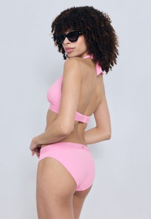 Femeie cu păr creț, purtând ochelari de soare negri și un costum de baie în două piese roz cu bretele la gât, pozând cu spatele la un fundal simplu și deschis la culoare.