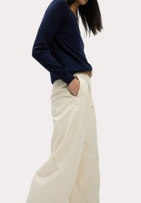 Pull à col en V à manches longues navy associé à un pantalon large crème. Le pantalon présente des plis et des poches ; le pull a des poignets côtelés.