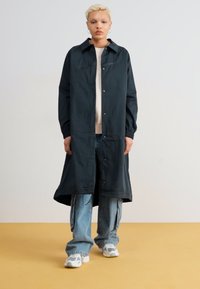 Dlhý čierny kabát s gombíkmi vpredu, strihaný na telo, s manžetami na rukávoch, v kombinácii s casual modrými džínsami a športovými topánkami.