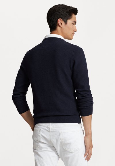 Sweater de malha azul marinho com mangas longas e punhos canelados, usado sobre uma camisa branca com colarinho. Apresenta um padrão de malha texturizada simples.