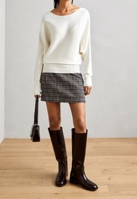 Witte geribbelde trui, zwart-wit gepatternde minirok, donkerbruin kniehoge laarzen, en een kleine zwarte handtas.