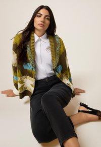 Neulejakku, jossa on vihreä, valkoinen ja sininen geometrinen kuvio, fringed-reunat, käytetty valkoisen paidan päällä ja yhdistetty mustien housujen ja korkokenkien kanssa.