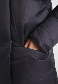 Marineblå puffjakke med synlig sidelomme. Materialet virker glat og lidt skinnende, med quiltet tekstur og bløde kanter.