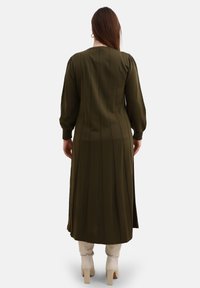 Olivenfarbenes Kleid mit langen Ärmeln und vertikalen Falten, mit einem runden Ausschnitt und einem bodenlangen Design, kombiniert mit weißen Stiefeln.
