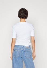 Valkoinen ribattua kangasta oleva lyhythihainen toppi, jossa on istuva malli, yhdistettynä korkeavyötäröisiin vaaleansinisiin farkkuihin, joissa on takataskut ja brändilogo.