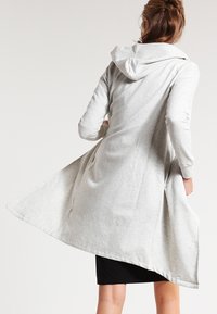 Cárdigan largo gris claro con capucha, que presenta mangas largas y un frente abierto. Está confeccionado con una tela suave y texturizada con un diseño fluido.