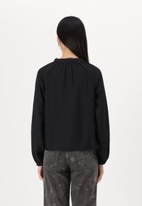 Svart långärmad topp med rundad halsringning och rynkad detalj, tillverkad av lättviktsmaterial. Matchas med grå jeans med stjärnmönster.