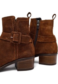 Stivaletti in suede marrone fino alla caviglia, con punta arrotondata, chiusura con zip laterale, fibbia decorativa e tacco in legno impilato. Presentano dettagli di cucitura visibili.