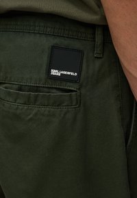 Vaqueros verde oliva con una etiqueta negra rectangular que dice "KARL LAGERFELD JEANS" en texto blanco, detalle visible del bolsillo trasero, textura suave.