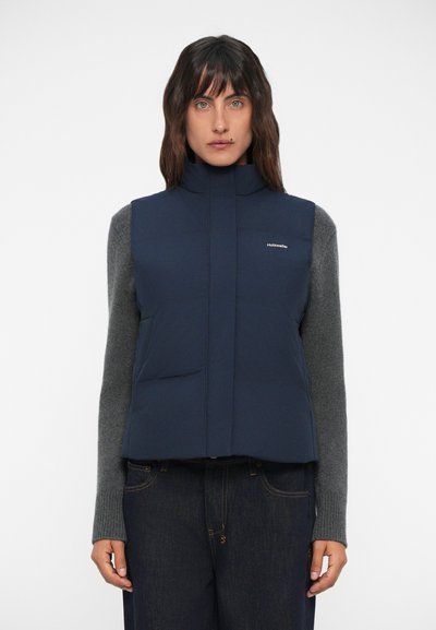 Gilet isolé bleu marine avec col haut et poches avant, associé à un pull gris à manches longues et un jean foncé. Design simple et moderne.