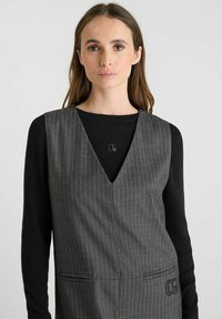 Grijs gestreepte vest met V-hals ontwerp en twee voorzakken, gelaagd over een zwart geribd lange mouw top. Bevat een logo-accent.