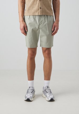 Pantalones cortos verde claro hechos de una tela suave y mate, con una cinturilla elástica. Llevados con zapatillas deportivas grises y calcetines blancos.