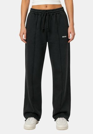 Persona che indossa pantaloni della tuta neri con coulisse e cuciture verticali e scarpe da ginnastica bianche, mostrata dalla vita ai piedi.