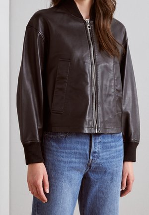 Femme portant une veste bombardier en cuir brun foncé avec fermeture éclair, poignets et taille côtelés, associée à un jean en denim bleu.