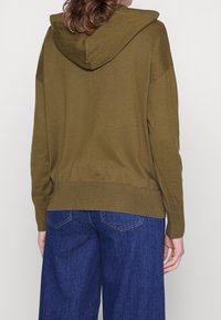 Oliven grøn hoodie med en afslappet pasform, ribbet kant og manchetter samt en stor hætte. Kombineret med brede blå denim bukser.