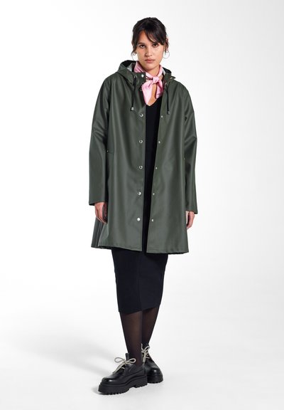 Imperméable vert avec capuche, boutons-pression et cordons de serrage, porté sur une robe noire et associé à de chics chaussures à plateforme noires.