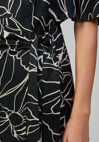 Robe en tissu noir ornée d'un motif floral blanc, mise en valeur par un détail de taille nouée. Texture douce avec des lignes de design fluides.