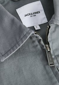 Gros plan d'une veste en tissu gris avec col, tirette de fermeture éclair visible et étiquette indiquant la marque Jack & Jones et la taille L.